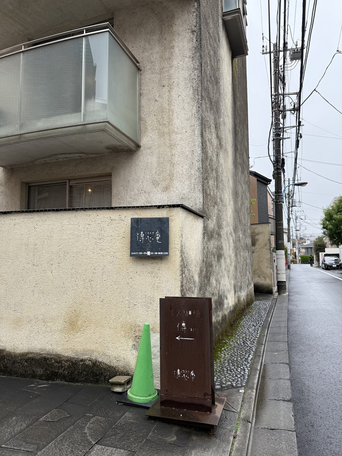 住宅街の路地に置かれた錆びた茶色の立て看板に「大火珈琲 傳」と白字で書かれ、左矢印と店名が手書きされている。背後にコンクリート壁と「傳心庵」の黒いプレートサイン