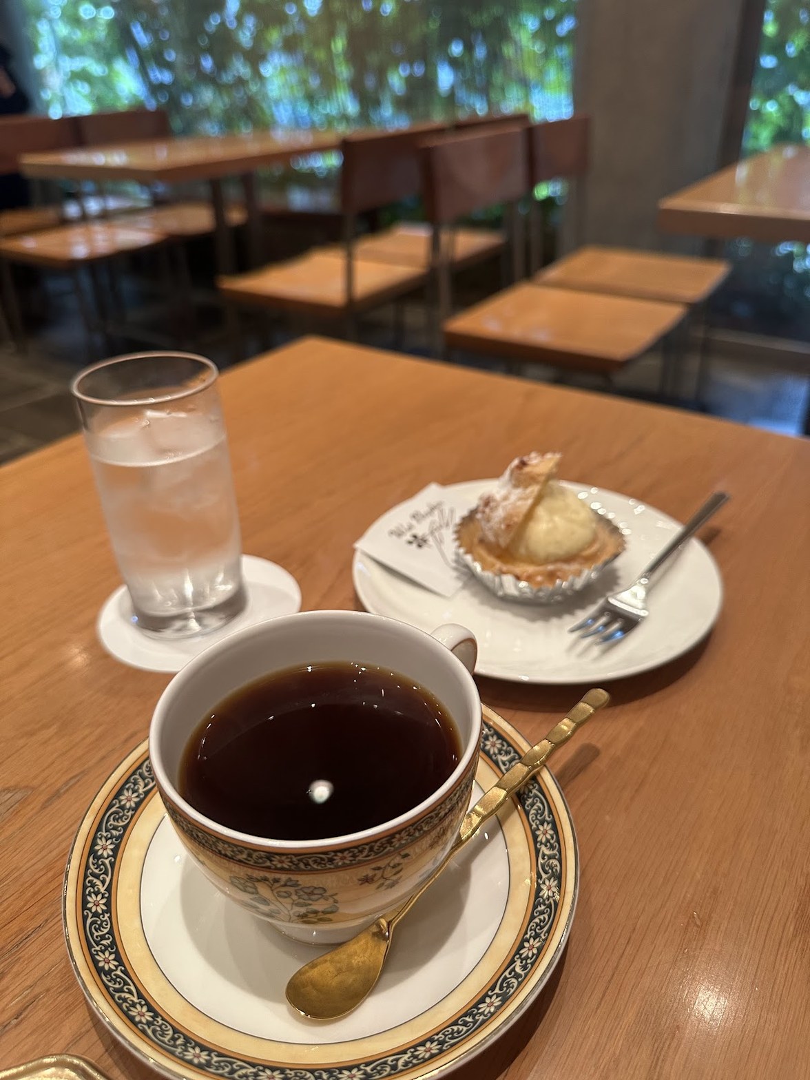 木製テーブルに置かれた金縁の白磁カップに注がれたブラックコーヒーと、奥にシュークリームの皿・水のグラス。背後に緑の木々が透けるガラス窓と空いた木製テーブル席が並ぶ