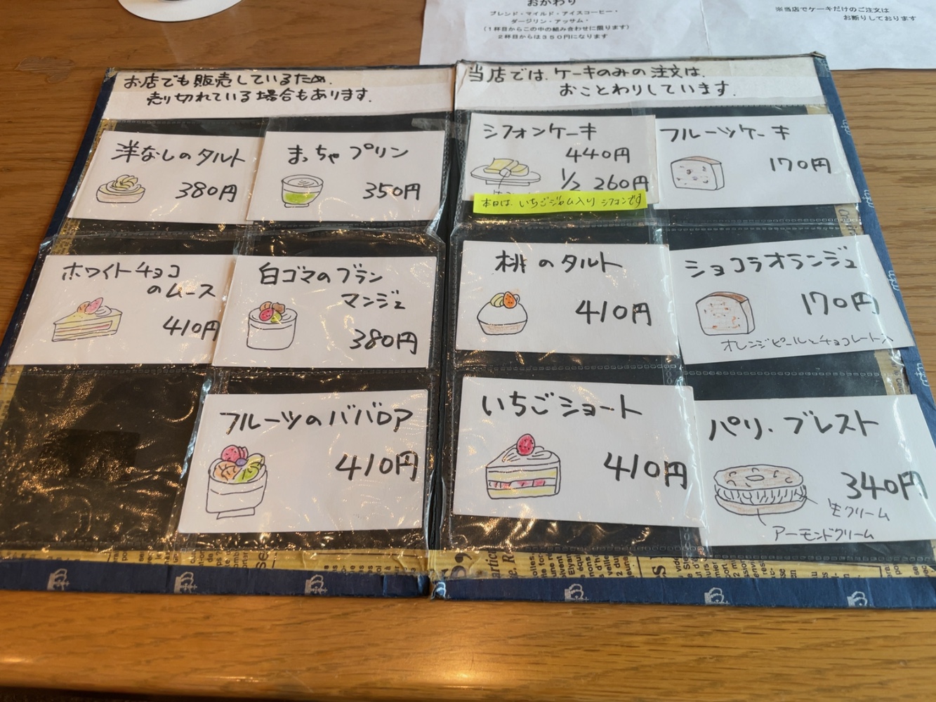 テーブルに広げられた手書きイラスト付きのケーキメニュー表。洋なしのタルト380円、シフォンケーキ440円、いちごショート410円など全品目と価格が記載されている