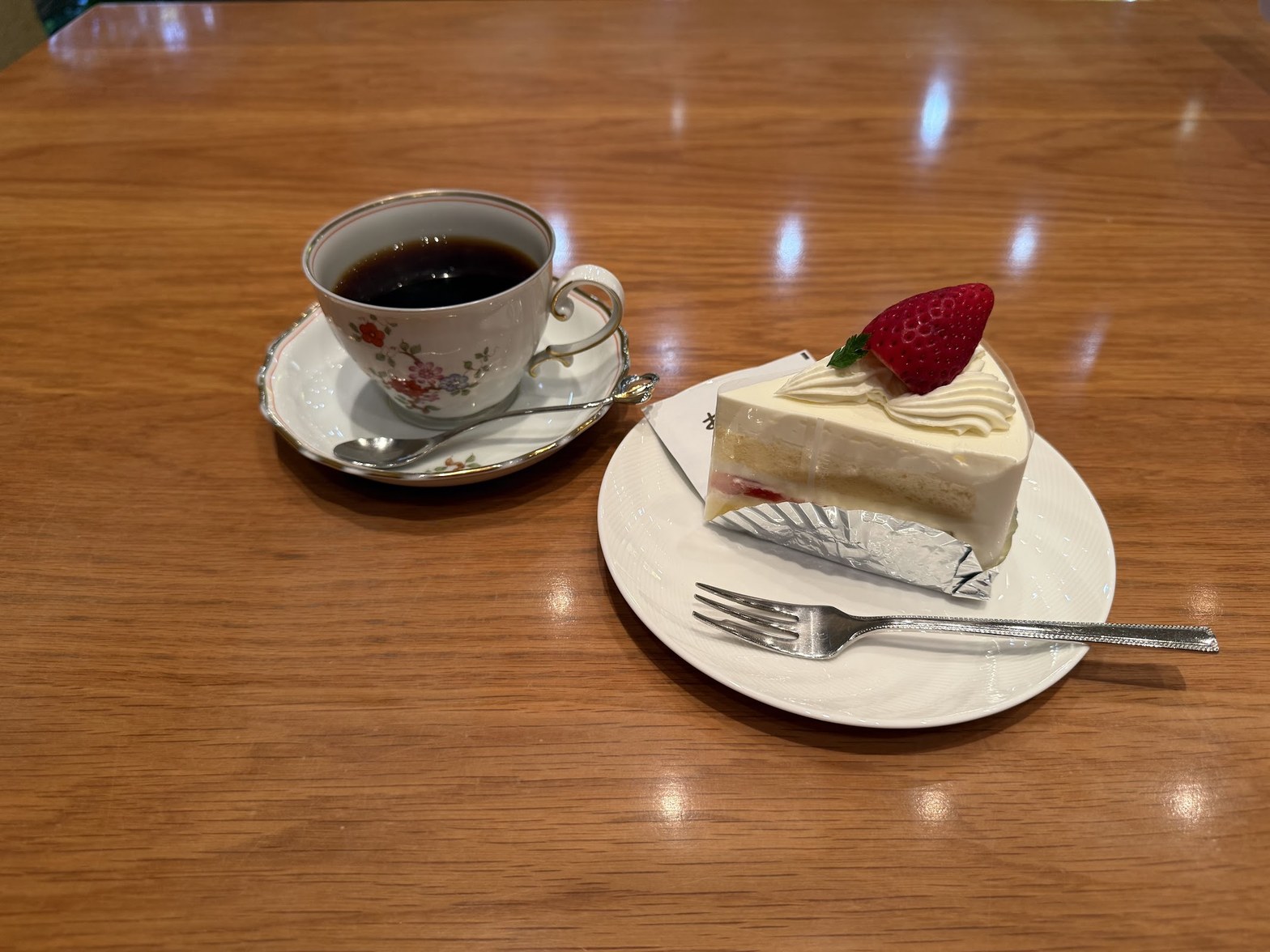木製テーブルの上に白磁の花柄カップに入ったブラックコーヒーと、いちごをのせたショートケーキの白い皿が並ぶ